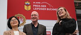 Foto: Die drei Preisträger*innen Ki-Hyang Lee, Tom Holert und Barbi Marković stehen lachend auf der Leipziger Buchmesse. Ki-Hyang Lee hält einen Blumenstrauß. Im Hintergrund steht auf der Wand geschrieben: "20 Jahre Preis der Leipziger Buchmesse"