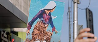 Ein Plakat: Eine Palästinensische Frau in traditioneller Kleidung erhebt sich aus den Trümmern Gazas