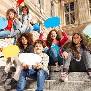 Kinder sitzen auf einer Treppe vor einer Schule und halten Schilder in Form von Sprechblasen hoch 