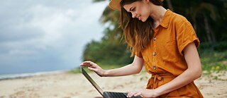 Eine junge Frau sitzt im Sand am Strand und schaut lächelnd auf ihren Laptop, sie trägt Sommerkleid und einen Hut