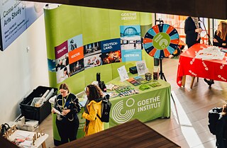 Blick von oben auf einen Veranstaltungsraum mit Ständen und mehreren Menschen 