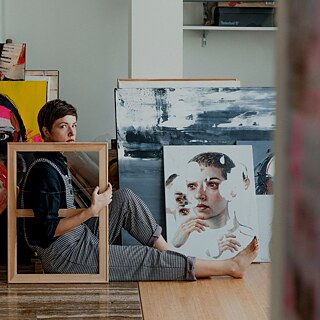 An artist sits in front of her paintings in her studio. She holds an empty picture frame in front of her body and looks seriously into the camera.
