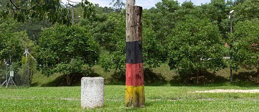 A foto de autoria de Bruna Engel mostra um poste de madeira pintado com as cores preto, vermelho e amarelo, ao lado de um pilar de concreto branco, ambos situados em um campo gramado com árvores ao fundo.