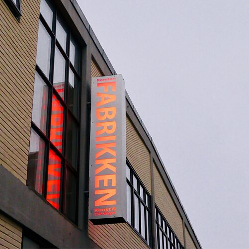 På billedet ses bygningen til FABRIKKEN for Kunst og Design udefra, på et neonskilte står FABRIKKEN for Kunst og Design, skriften spejler seig i et af vinduerne.