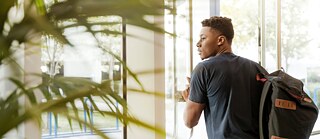 Ein junger Mann mit einem Rücksack verlässt ein Gebäude durch eine Glastüre.