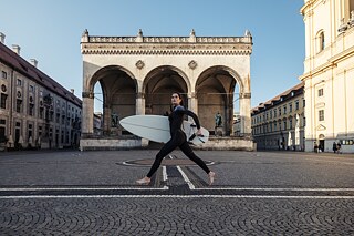 Eine Frau, die ein Surfboard trägt läuft über einen Platz in der Innenstadt