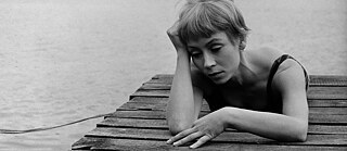 A woman with short blond her and a melanscholic expression is lying on a jetty  by a lake