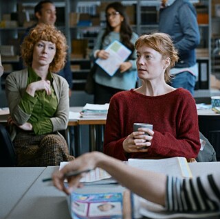 Eine Frau, sitzend, einen Becher haltend sitzt im Zentrum eines Raums, vor und neben ihr und im Hintergrund weitere Personen (Das Lehrerzimmer)