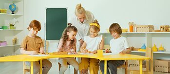 Schüler:innen der Grundschule sitzen an einem Tisch. Ihre Lehrerein steht daneben und schaut auf ihre Arbeiten