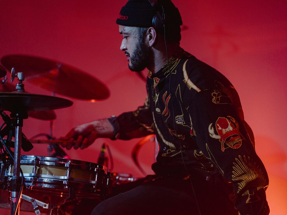 Male with patterned shirt plays the drums under red lighting  