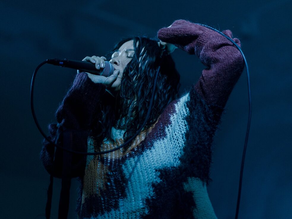 Felicia sings into a microphone with arm raised under blue lighting 