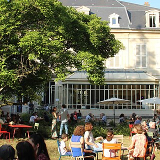 La photo montre des personnes dans un jardin en été. En arrière-plan, on aperçoit le Goethe-Institut entre les grands arbres.