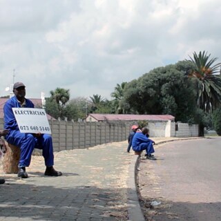 People sitting next to a street, holding up placards, seeking work