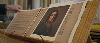 close up photo of a music stand on top of piano with a volume of music, an image of a women in 19th century hair style