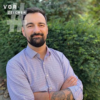 Das Bild zeigt Ervin Malakaj, ein Mann in einem karierten Hemd mit Bart, der vor einer Hecke steht. Links oben im Bild ist das Logo der Reihe Vorzeichen zu sehen.