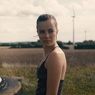 a young woman standing in a field, wind turbines in the background