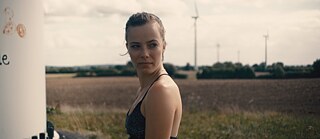 a young woman standing in a field, wind turbines in the background