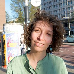 The picture shows Leila Essa, a woman with brown, curly hair, wearing a green blouse. The picture was taken outside on the street and in the background you can see a house, a railway station clock and posters.