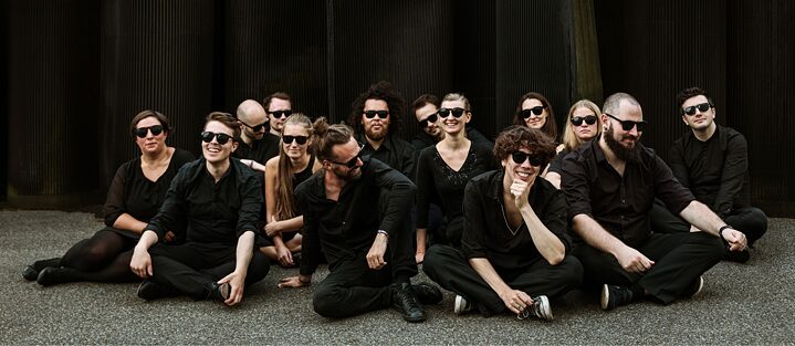 Die Mitglieder des Orchester im Treppenhaus sitzen draußen auf dem Boden, alle tragen schwarze Kleidung und schwarze Sonnenbrillen. 