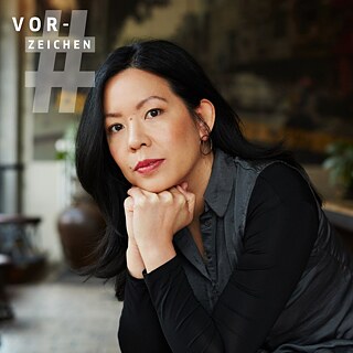 Auf dem Bild ist die Autorin Khuê Phạm zu sehen, eine Frau mit langen, schwarzen Haaren. Sie stützt das Gesicht in den Händen ab und schaut direkt in die Kamera.