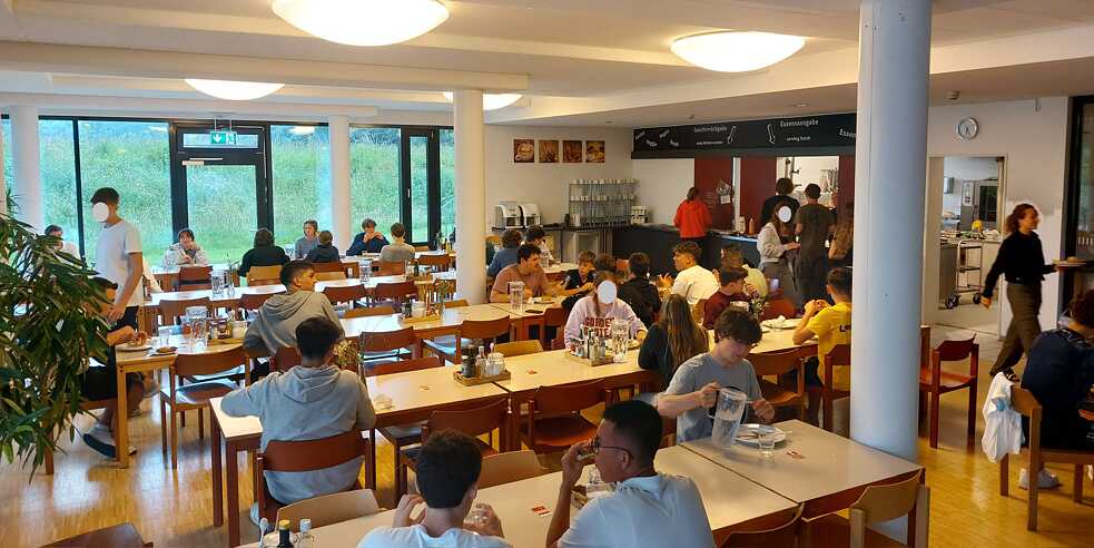 Eine große, helle Kantine, modern eingerichtet. Jugendliche sitzen an den Tischen und genießen ihre Speisen.