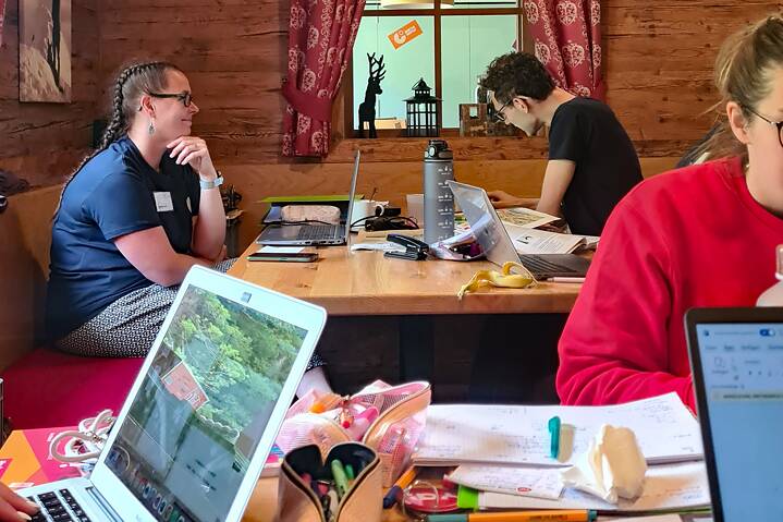 Junge Menschen in einer Blockhütte sitzen an Tischen. Vor ihnen liegen Laptops und Schreibuntensilien.