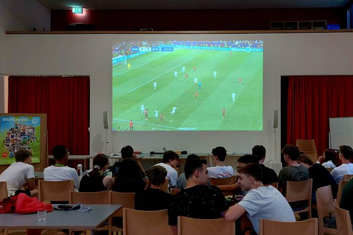 In einem Raum hängt eine große Leinwand, darauf wird ein Fußballspiel projeziert. Jugendliche sehen angespannt zu und unterhalten sich.