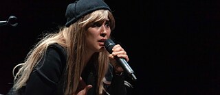 Eine Frau performt auf einer dunklen Bühne mit Mikrofon.