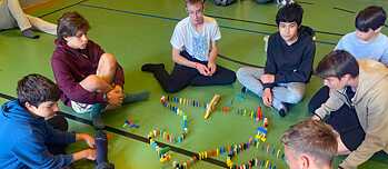 Jugendliche sitzen auf dem Fußboden einer Turnhalle und stellen bunte Dominosteine zu einer Kette auf.