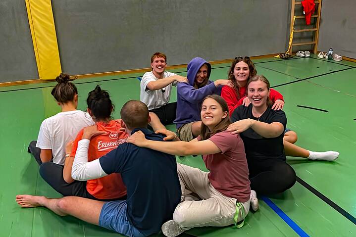 Eine Gruppe Jugendlicher sitzt in einem Kreis auf dem Fußboden einer Sporthalle und lächtelt in die Kamera.