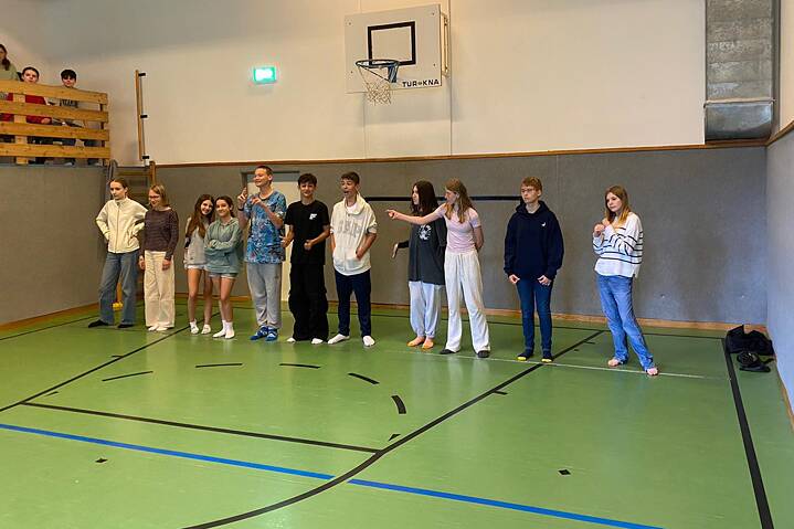 Eine Gruppe Jugendlicher steht nebeneinander am Kopfende einer Sporthalle. Über ihnen hängt ein Basketballkorb.