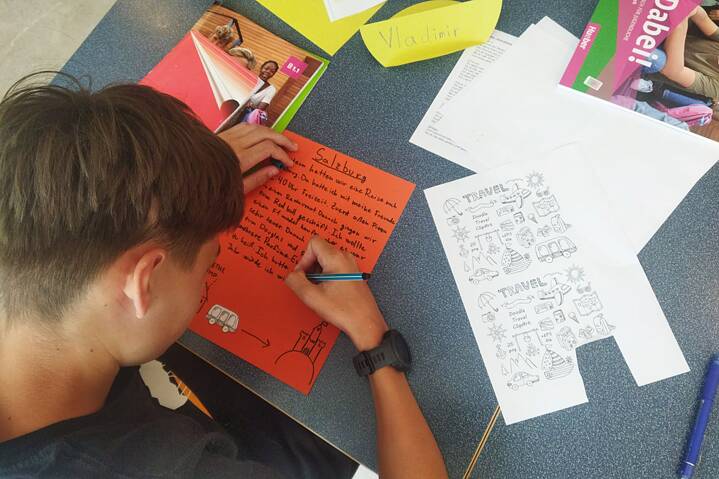 Ein Jugendlicher sitzt an einem Tisch und schreibt auf einem orangefarbenen Papier.