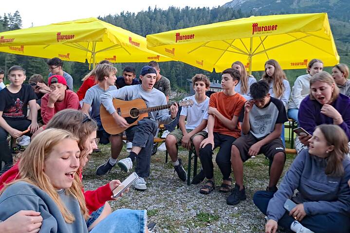 Jugendliche sitzen unter großen Sonnenschirmen, einer spielt Gitarre, alle anderen singen.