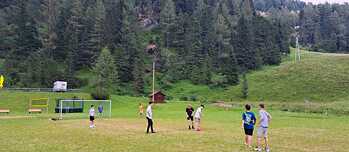 Jugendliche Spielen Fußball auf einer Wiese; im Hintergrund ist ein Wald zu sehen.