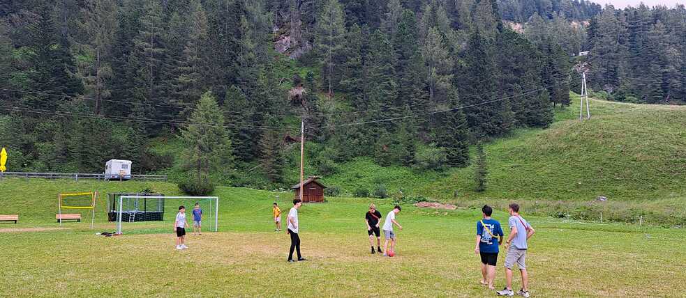 Jugendliche Spielen Fußball auf einer Wiese; im Hintergrund ist ein Wald zu sehen.