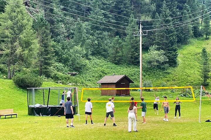 Jugendliche spielen Volleyball auf einer Wiese. Im Hitnergrund sieht man bewaldete Berge.