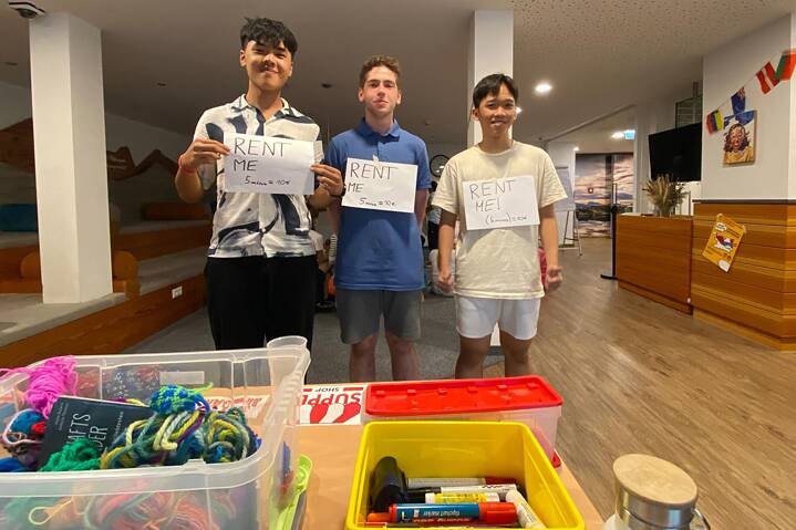 Drei Jugendliche halten ein Schild, auf dem steht: „Rent me. 10€“. Im Vordergrund sieht man Plastikkästen mit buntem Krimskrams.