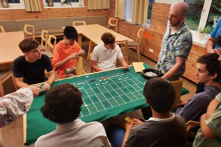 Jugendliche sitzen um einem grünen, mit Zahlen und schwarz/roten Feldern beschriebenen Tisch und spielen Roulette.