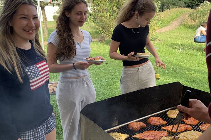 Drei junge Frauen stehen mit Tellern in der Hand vor einem Grill, auf dem mariniertes Fleisch liegt.