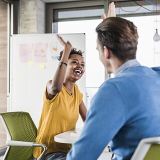 Zwei fröhliche junge Menschen klatschen die Hände gegeneinander und lachen. Im Hintergrund steht eine Whiteboard.