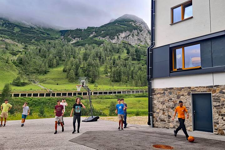 Jugendliche spielen Ball vor einer Herberge in den Alpen, im Hintergrund sind hohe, wolkenverhangene Berge zu sehen.