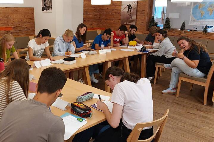 Jugendliche arbeiten konzentriert an einer Tischanordnung in L-Form. Eine junge Erwachsene freutlilch sich.