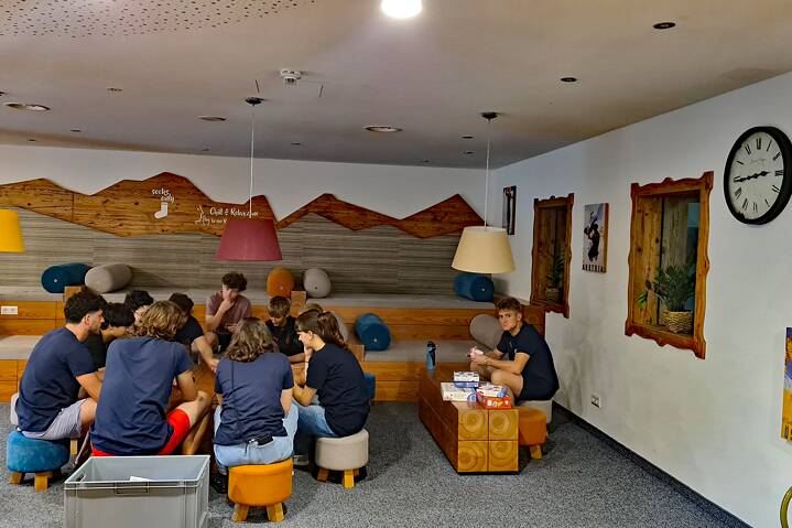 Jugendliche sitzen Zusammen auf niedrigen, bunten Hockern und spielen Brett-/Gesellschaftsspiele. Der Raum ist im alpinen Stil eingerichtet und dekoriert.