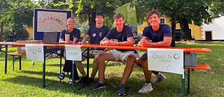 Junge Erwachsene sitzen an einem langen Biertisch und lächeln in die Kamera.