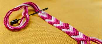 Auf einer gelben Decke ist ein Freundschaftsband in Pink und Weiß mit einer Sicherheitsnadel befestigt. Es ist noch nicht fertig geknäüft.