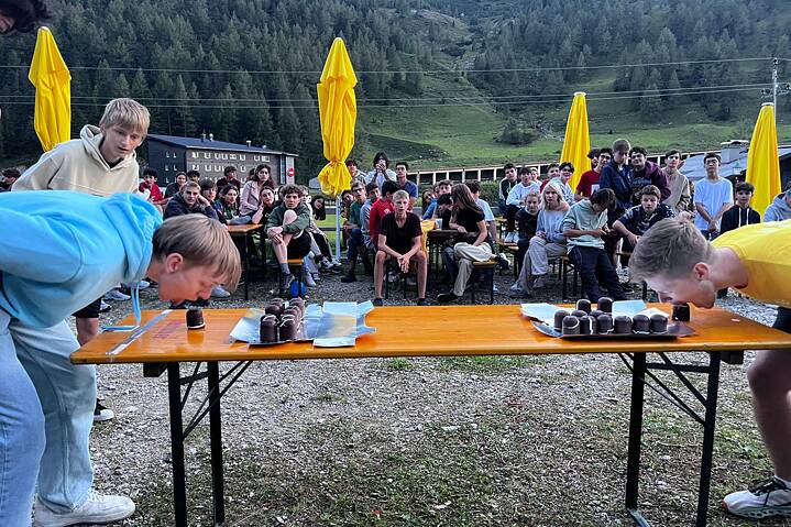 Auf einem langen Biertisch liegen schokoladige Schaumküsse. Jugendliche versuchen diese zu essen, ohne die Hände dabei zu benutzen.