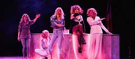 Five women do their performance on a theater stage in pink light.