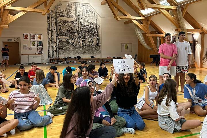 Jugendliche sitzen auf dem Boden und halten ein Plakat hoch, auf dem steht: „Wir lieben Euch“.