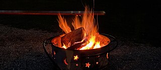 In einer Schale lodert ein Feuer, dahinter steht eine Bierbank.