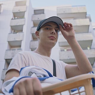 Junger Mann mit Schirmmütze und Wäschekorb vor Hochhausblock